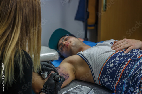 A boy with glasses and blue cap is tattooed by a young woman in her tattoo studio