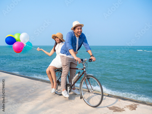Happy Asian couple riding bicycle and holding colorful balloons on the beach, lifestyle concept.