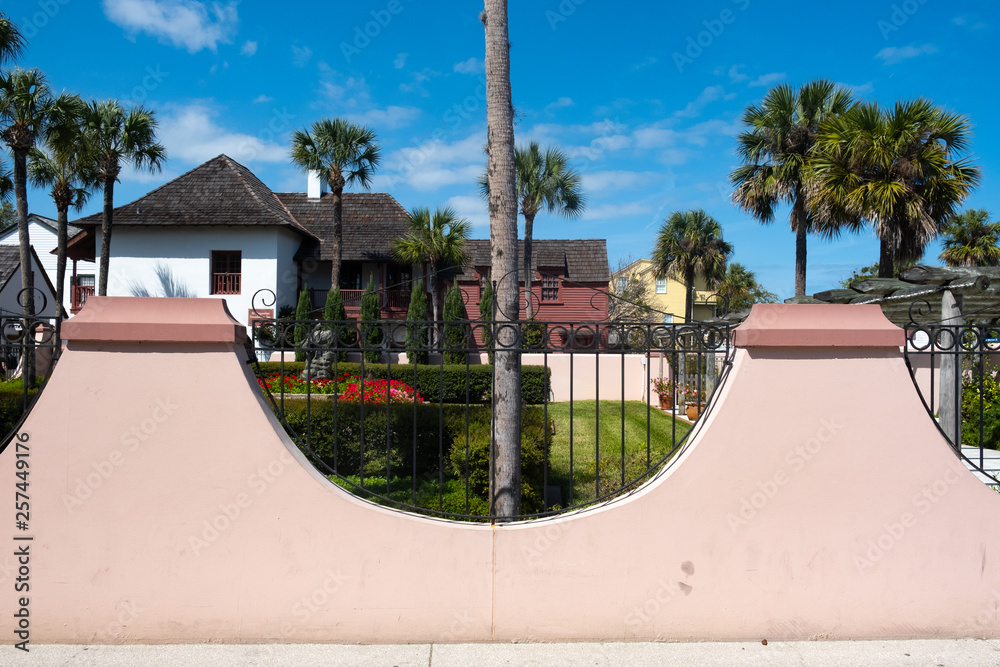 Obraz premium Half oval pink wall with wrought iron fence against a deep blue sky, palm trees and a home