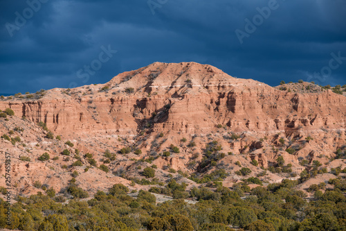 Bright sunlight illuminates a colorful red rock desert peak and badlands under dark, dramatic storm clouds near Santa Fe, New Mexico