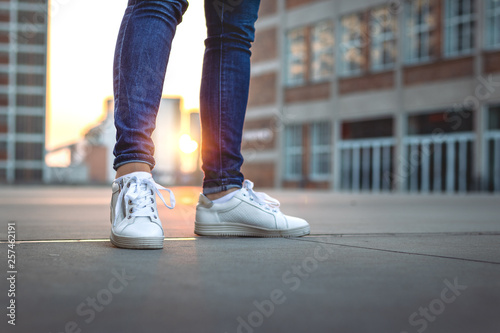 Woman wearing jeans and sneakers is standing in city during sunset. Selective focus