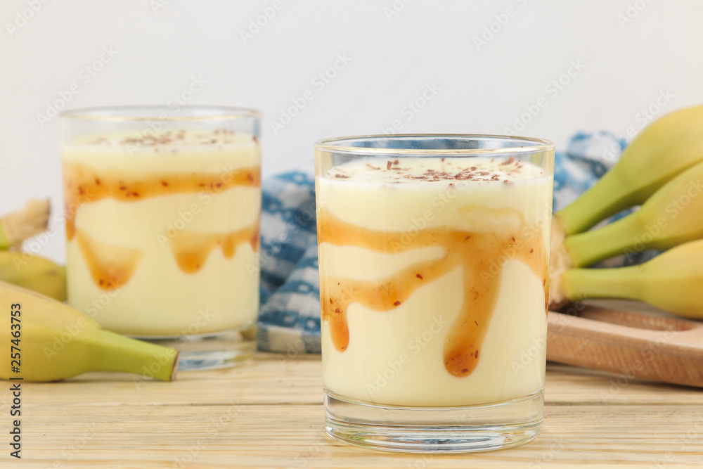 a glass with a banana milkshake, syrup and chocolate chips and a fresh banana on a natural wooden table. Making a milkshake. a glass with a banana milkshake, syrup and chocolate chips and a fresh banana on a natural wooden table. Making a milkshake.