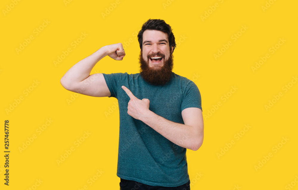 Handsome fit young funny bearded man pointing to his bicep and smiling on yellow background. Handsome fit young funny bearded man pointing to his bicep and smiling on yellow background.