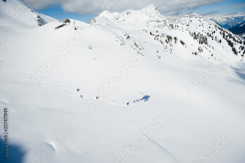 A group of skiers follow thier guide down a large alpine bowl in the Selkirk Mountains, Canada while the shadow of their helicopter is follows. photo