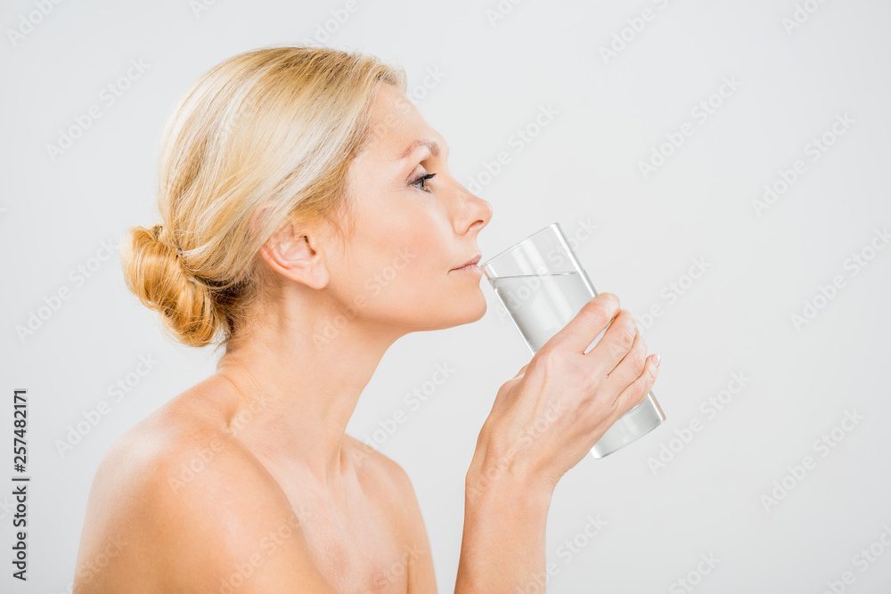 side view of mature woman drinking water from glass isolated on grey side view of mature woman drinking water from glass isolated on grey