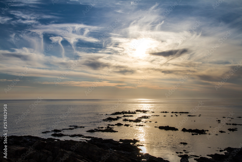 Obraz premium Blue sky at dawn covered by strange cloud formations on a flat sea with black rocks that stand out in the reflection of the sun on the water