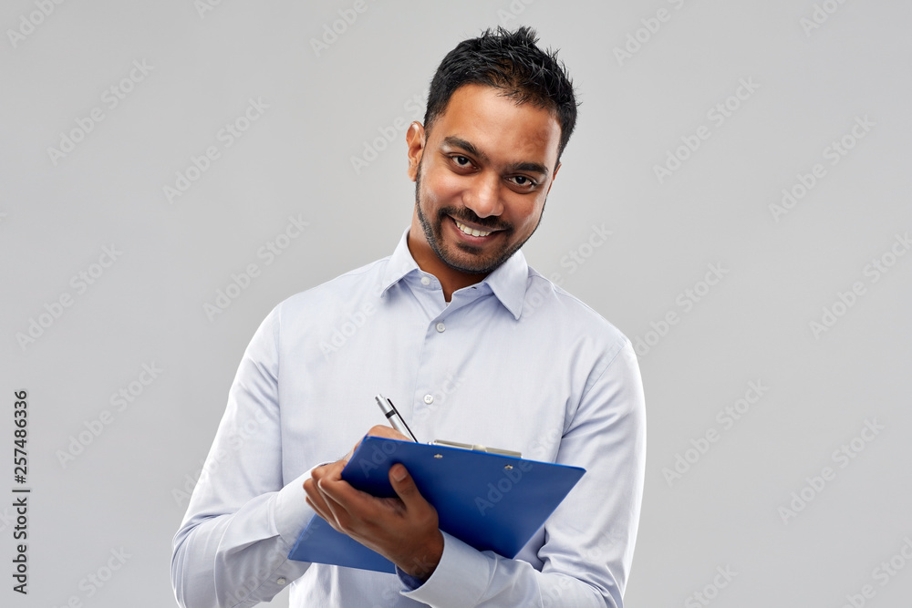 business, office worker and people concept - smiling indian businessman writing to clipboard over grey background business, office worker and people concept - smiling indian businessman writing to clipboard over grey background