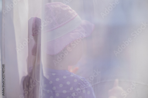 little toddler girl behind curtains looking out of the window in sunny weather, sad lonely child abuse family domestic violence theme concept, melancholy, depression, home alone, escape.window