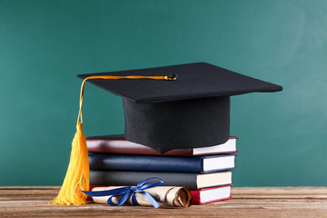 Graduation cap with diploma and stack of books on green board background