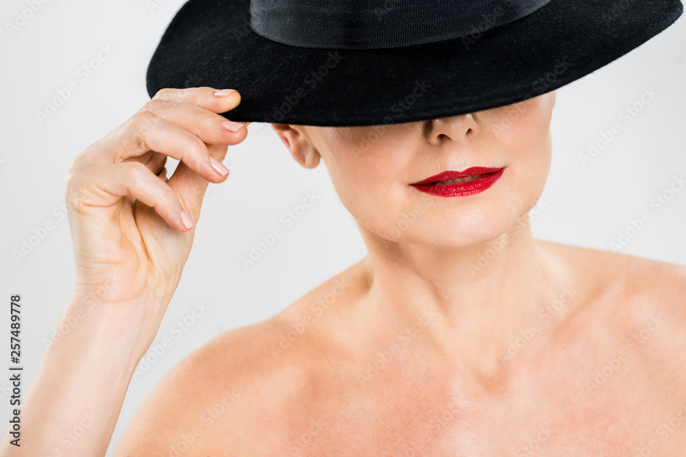 middle aged elegant and fashionable woman with red lips touching black hat isolated on grey middle aged elegant and fashionable woman with red lips touching black hat isolated on grey