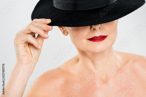 middle aged elegant and fashionable woman with red lips touching black hat isolated on grey