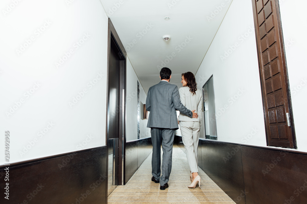 back view of confident businessman hugging businesswoman in formal wear while walking in hotel corridor back view of confident businessman hugging businesswoman in formal wear while walking in hotel corridor