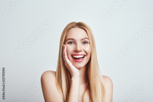 Make up for her. Cheerful young woman smiling happily, looking at camera and touching her chin while standing against abstract creative background