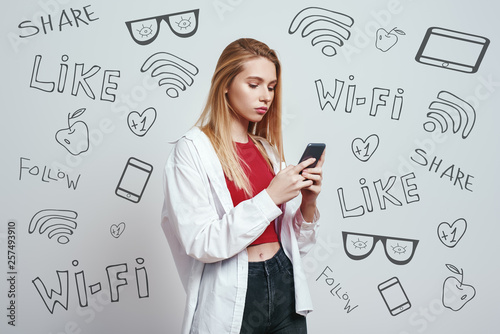 Surfing the net. Young blonde woman in a casual wear using her smart phone while standing against grey background with different doodle illustrations on it. Social media