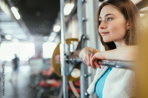 Young woman exercising in gym room. Nice cheerful model lean to barbell in smith machine and smile. Resting. Towel around neck. Alone in fitness room.