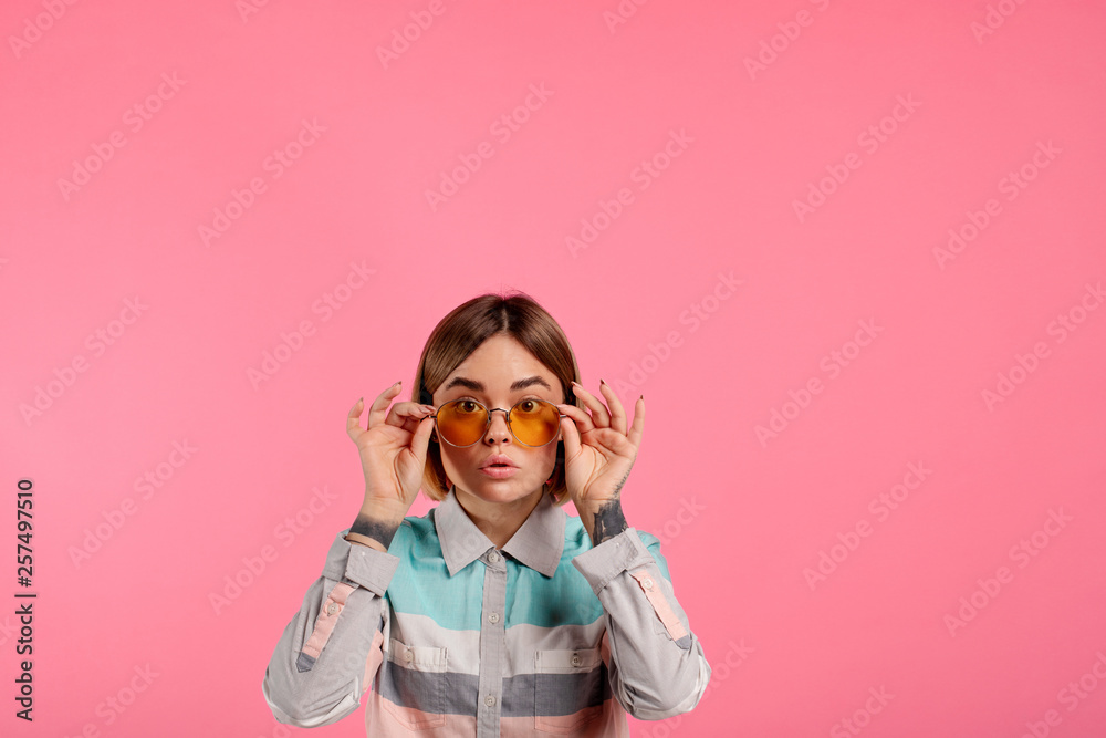 Beautiful fashioned young woman pose on camera. She hold sunglasses with hands and look straight. Serious and concentrated. Isolated over pink background.