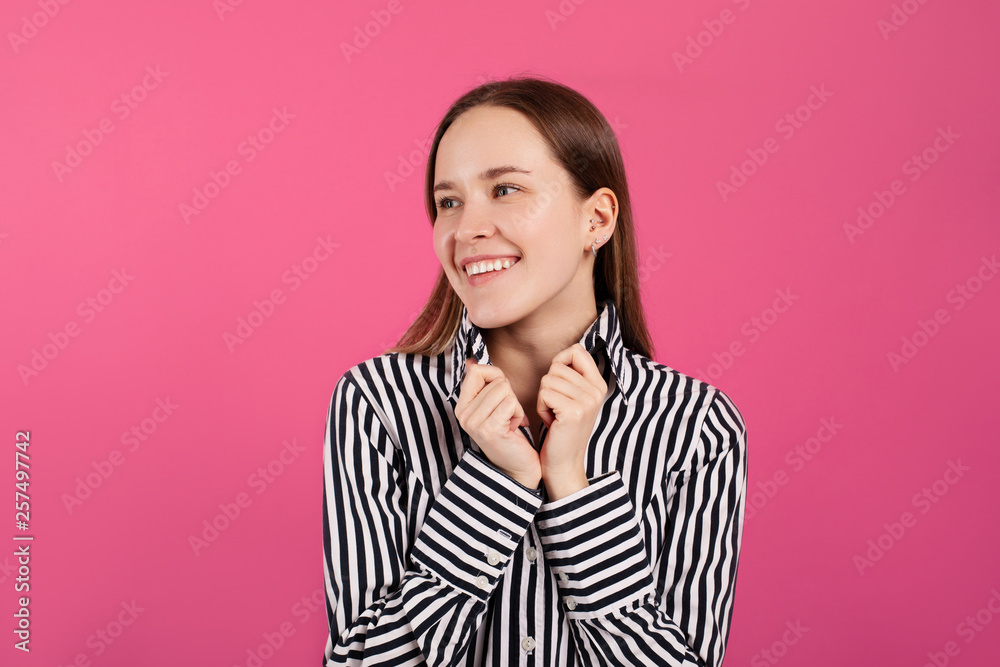 Cheerful nice brunette in striped shirt pose on camera and smile. She hold hands on collar and look to left. Isolated over dark pink background.