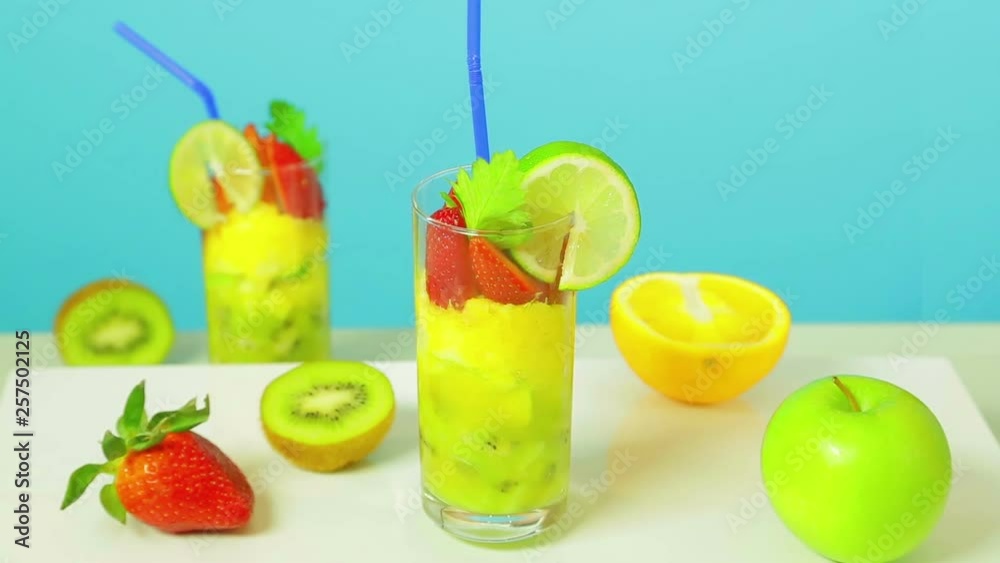 Fruit cocktail in a glass of kiwi, apple and strawberry slices decorated with lime with a blue straw on a blue background. Glass rotates in a circle. Fruit cocktail in a glass of kiwi, apple and strawberry slices decorated with lime with a blue straw on a blue background. Glass rotates in a circle.