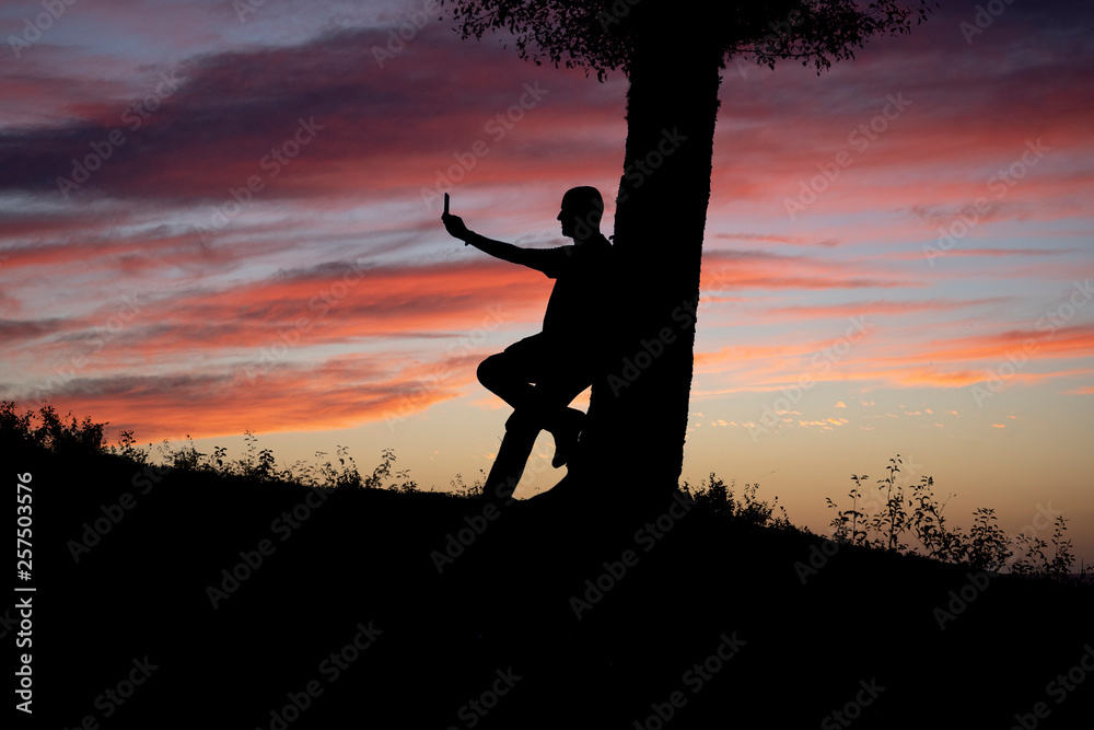 silhouette of a man photographing the landscape during sunset with his mobile phone.