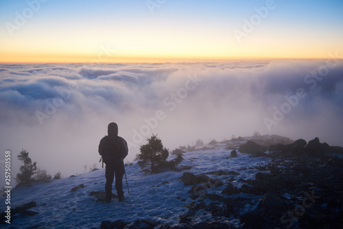 Back view of tourist with backpack on rocky snowy mountain peak on background of valley covered with white clouds stretching to horizon under bright morning sky with light orange glow at sunrise.