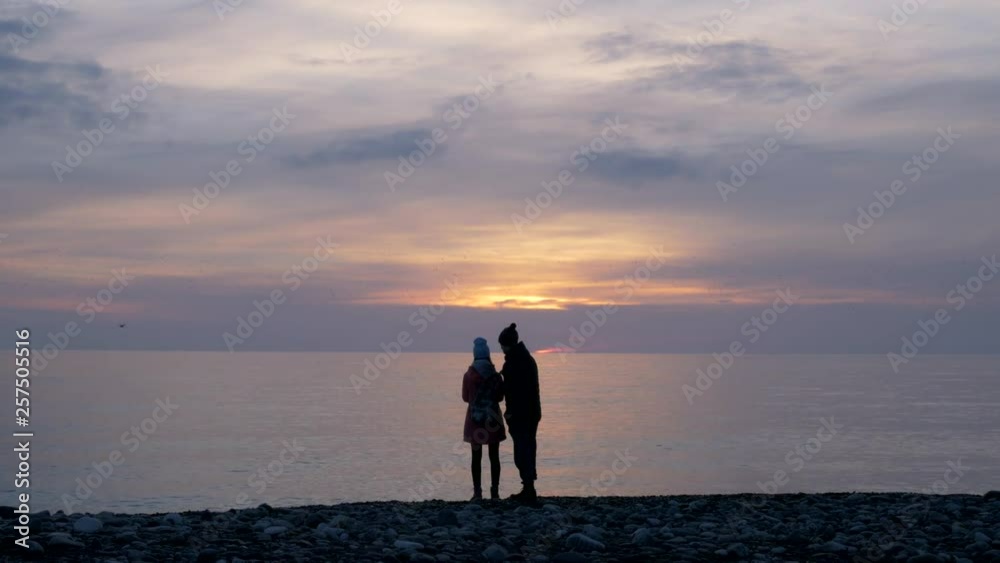 Young couple stands on the pebble stone sea beach and looks to sea during sunset. boy and girl in hats, cold weather, Flock of birds fly in sky. Couple enjoy of sunset Young couple stands on the pebble stone sea beach and looks to sea during sunset. boy and girl in hats, cold weather, Flock of birds fly in sky. Couple enjoy of sunset