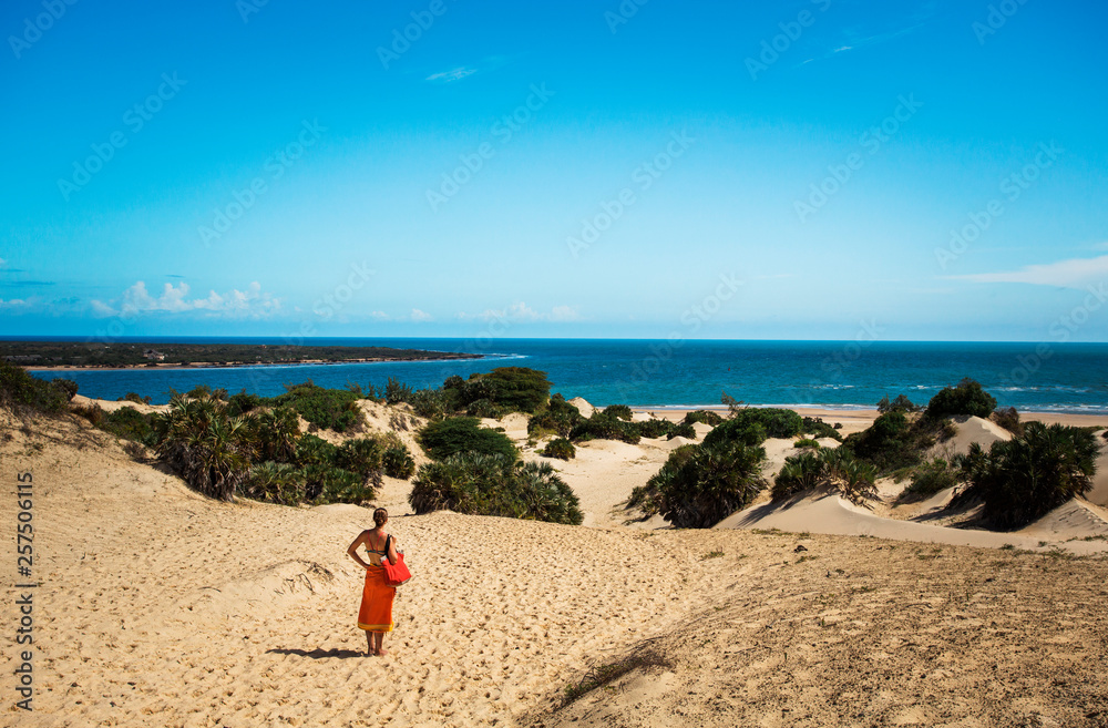 LAMU, INDIAN OCEAN, KENYA, AFRICA. A young woman in a bright orange sarong stands at the top of a vast landscape of dunes and blue water. LAMU, INDIAN OCEAN, KENYA, AFRICA. A young woman in a bright orange sarong stands at the top of a vast landscape of dunes and blue water.