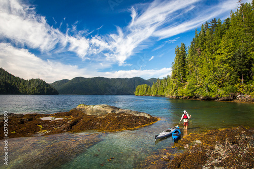 HAIDA GWAII, BRITISH COLUMBIA, CANADA. A man pulls two kayaks through shallow, turquoise water along a rocky, forested shoreline. photo