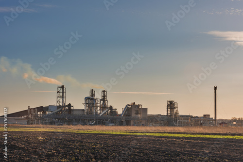 Industrial spring landscape with a view of a wood processing plant with high smoking pipes, against a background of blue sky and agricultural fields.