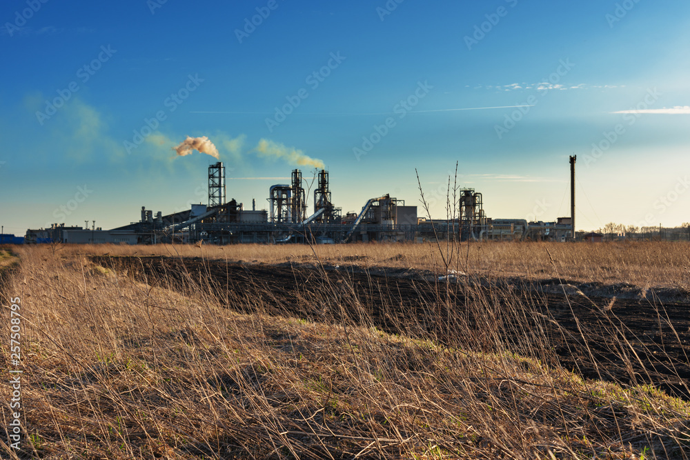 Fototapeta premium Industrial spring landscape with a view of a wood processing plant with high smoking pipes, against a background of blue sky and agricultural fields.