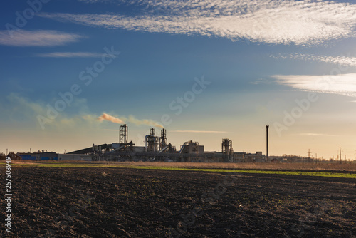 Industrial spring landscape with a view of a wood processing plant with high smoking pipes, against a background of blue sky and agricultural fields.