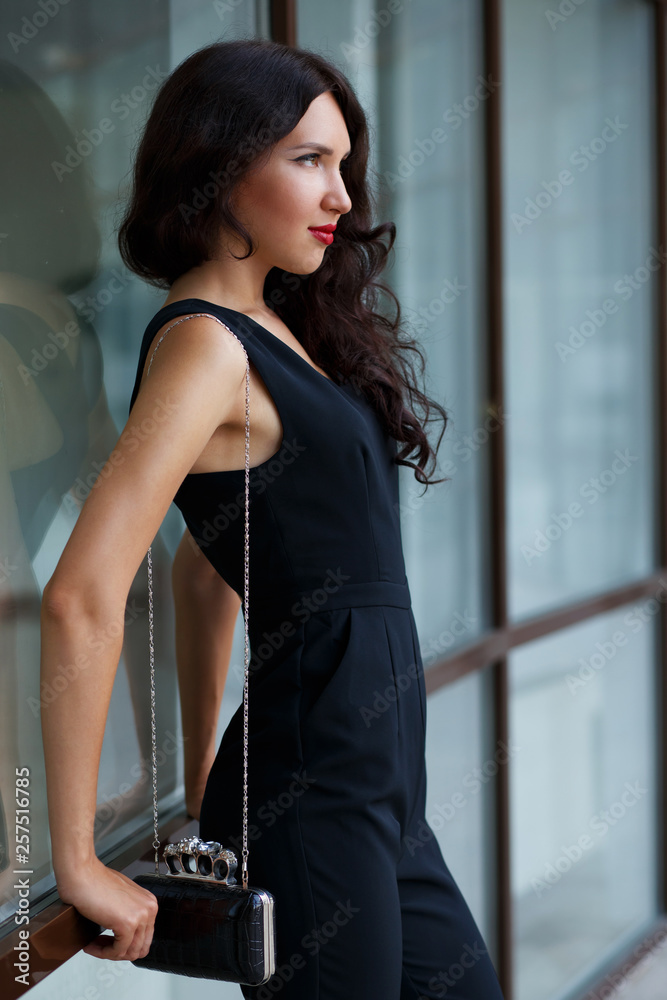 Beautiful young brunette woman with tanned skin, curly hair and red lips, wearing black jumpsuit and clutch bag on chain, posing outdoors near glass office building. Street fashion. Stylish outfit.