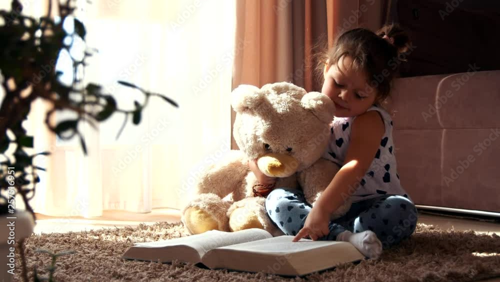 Little girl sitting on the floor reading a book hugging toy teddy bear ...