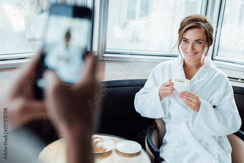cropped view of man taking photo of attractive girlfriend holding cup while sitting in bathrobe in hotel