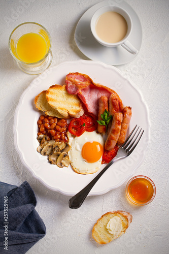 English or Irish breakfast in white plate with sausages, bacon, eggs, tomatoes, mushrooms, toasts and beans. vertical, overhead