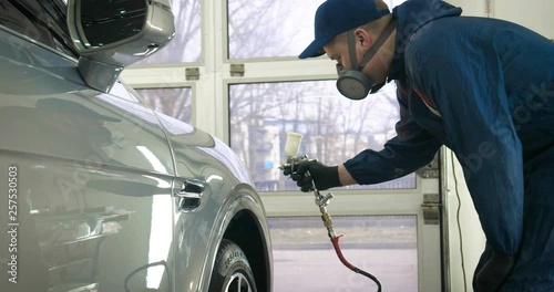 In slow motion, a Man (Man) professional in working uniform with spray guns in his hands and a respirator in protective glasses puts paint (ceramics) on the car on different parts of the car.