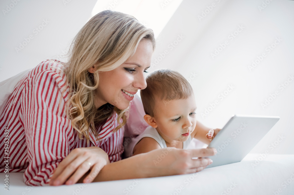 Smiling mother and toddler son lying in bed at home using tablet Smiling mother and toddler son lying in bed at home using tablet