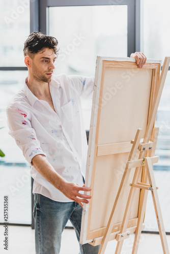 handsome artist in white shirt fixing canvas on easel in painting studio