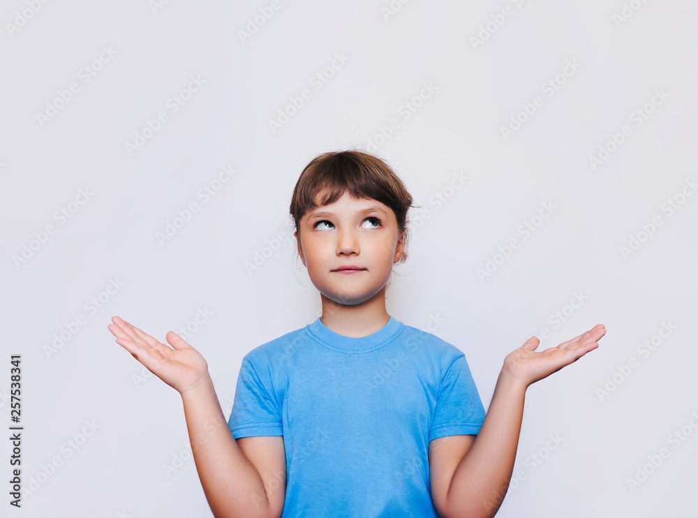 The little cute girl looks up in surprise and throws up her hands, as if pointing at something and expressing uncertainty. White background, copy space. The little cute girl looks up in surprise and throws up her hands, as if pointing at something and expressing uncertainty. White background, copy space.