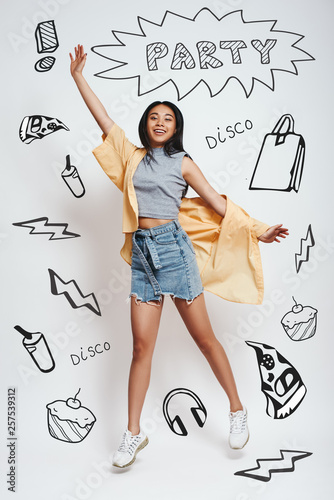 Going crazy! Happyyoung asian woman smiling and jumping against grey background with party theme doodles on it. photo