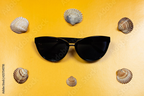 Flat lay na żółtym tle, okulary przeciwsłoneczne i muszelki. Inspiracja na wakacje, lato, czas wolny