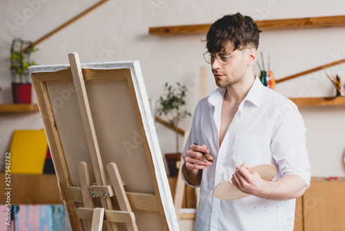 handsome artist in glasses standing at easel with canvas in painting studio