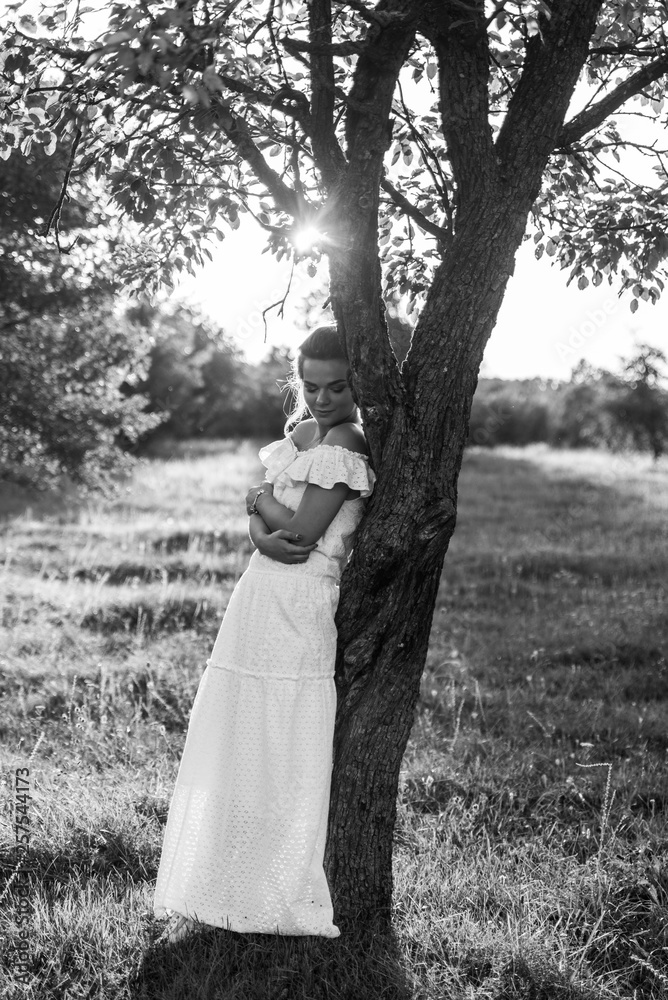 Awesome girl in a beautiful long dress posing in the garden near the tree. Black and white photo