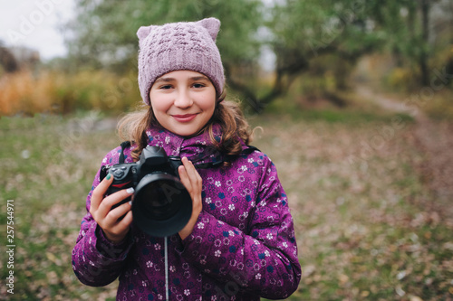 A young girl in a purple jacket and hat walks with a camera on the nature in the fall. Baby photographer. Happy childhood. Portrait and photography. © shchus