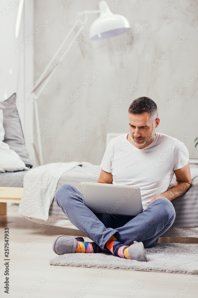 Handsome middle aged man sitting on the floor wirh crossed legs, leaning on bed and using laptop. Handsome middle aged man sitting on the floor wirh crossed legs, leaning on bed and using laptop.