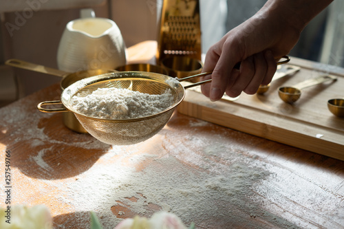 Flour sieve kept in a woman's hand. Rustic disorder reigns around you. On the table stands a flour, a dish with milk and spring tulips. The table is flecked with flour. Around are placed golden pots photo