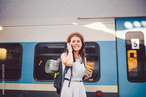 theme tourism and travel young student. beautiful young Caucasian girl in dress and hat standing at train station near train with backpack and cup coffee in hand. Uses smartphone phone technology