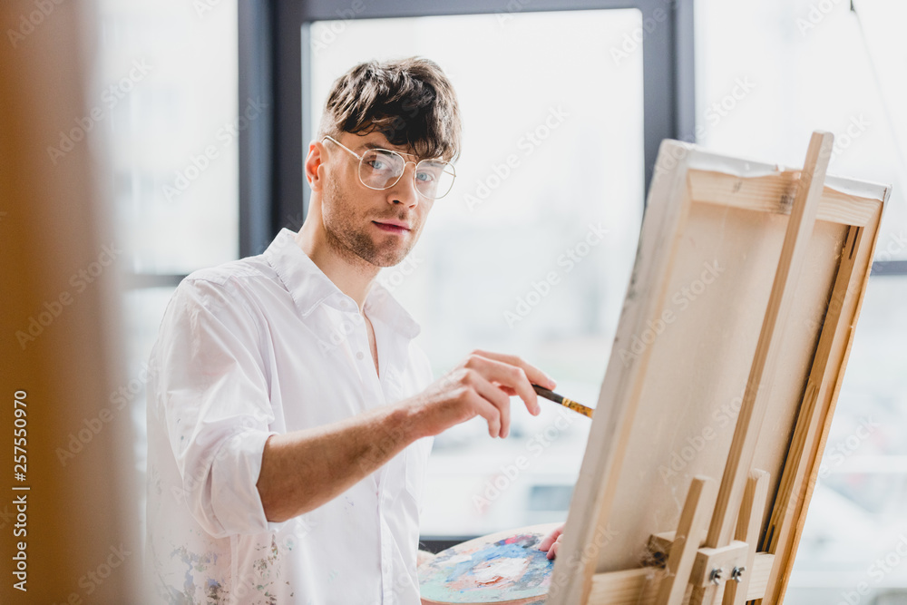 selective focus of handsome artist in glasses looking at camera while drawing on canvas