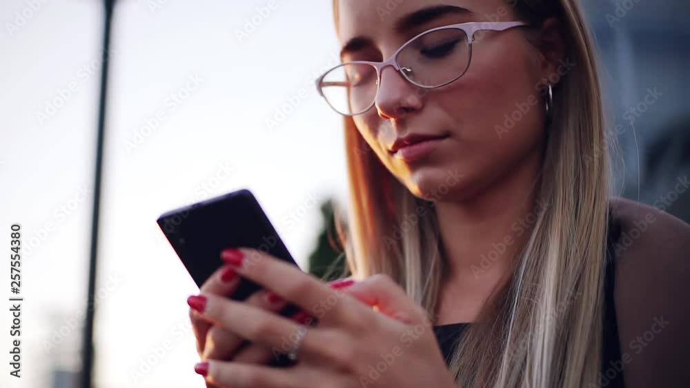 Young beautiful woman with red nails wear glasses use phone at sunset on a summer day smartphone girl holding mobile modern technology telephone texting touch using adult attractive looking message Young beautiful woman with red nails wear glasses use phone at sunset on a summer day smartphone girl holding mobile modern technology telephone texting touch using adult attractive looking message