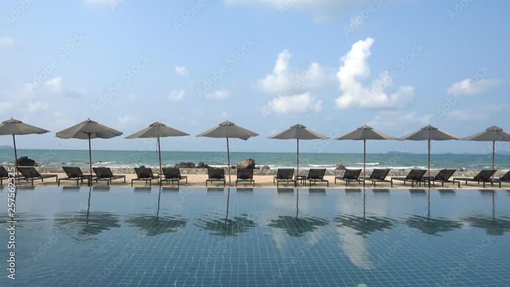 Along the edge of a resort pool sits multiple empty beach recliners shaded by umbrellas facing the incoming tide.
