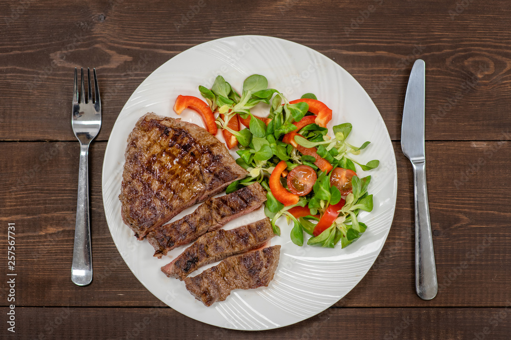 Grilled steak with vegetables on a plate with fork and knife on wooden background Grilled steak with vegetables on a plate with fork and knife on wooden background